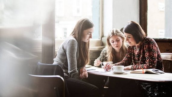 Three young women sat having coffee at a table and making notes