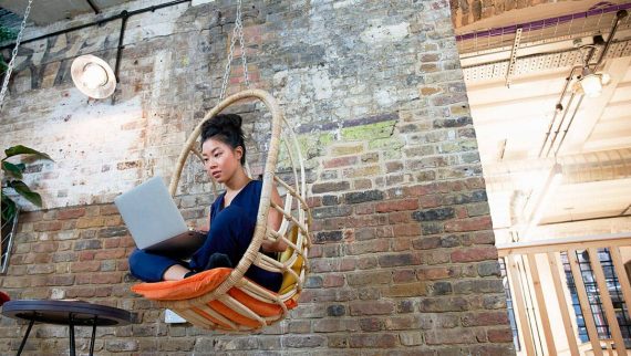 A young woman sat with her legs crossed outside on a chair swing looking at her laptop