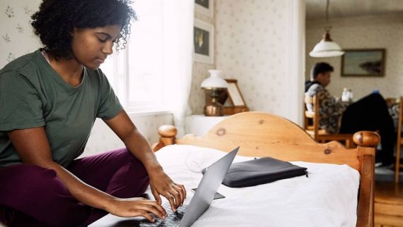A young woman sat on her bed typing on her laptop