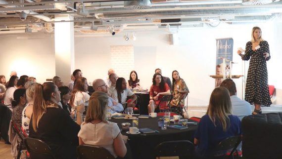 People at a roundtable event watching presenter speak on stage.